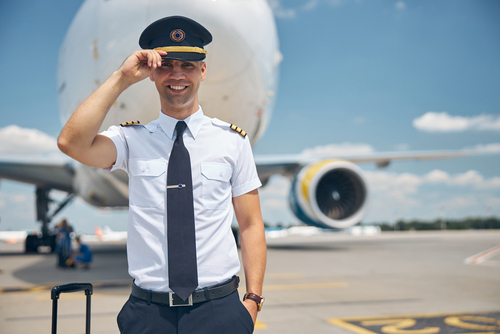 Jeune homme souriant et travailleur aérien touchant le chapeau du capitaine et souriant alors qu'il se tenait en terrain d'aviation avec un avion sur fond